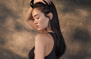 A woman with long dark hair in a black sleeveless top poses with one hand on her head against a textured background.